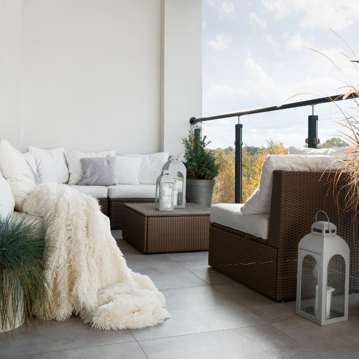 A balcony with comfy rattan sofa, throws, lanterns, and plants suggesting an inviting and cosy outdoor space.