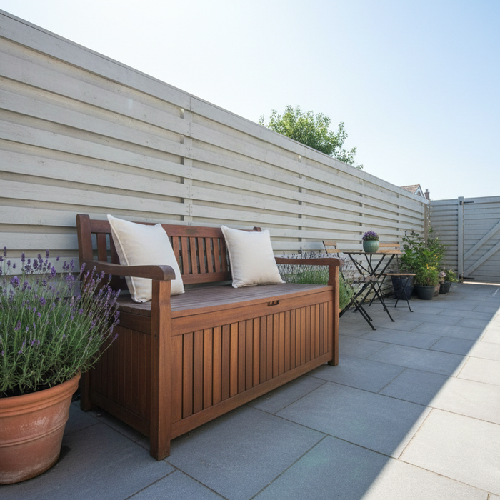 Wooden outdoor storage bench with neutral cushions on top, beside a terracotta pot of lavender, on a bright sunny UK patio with grey paving and a light grey fence.