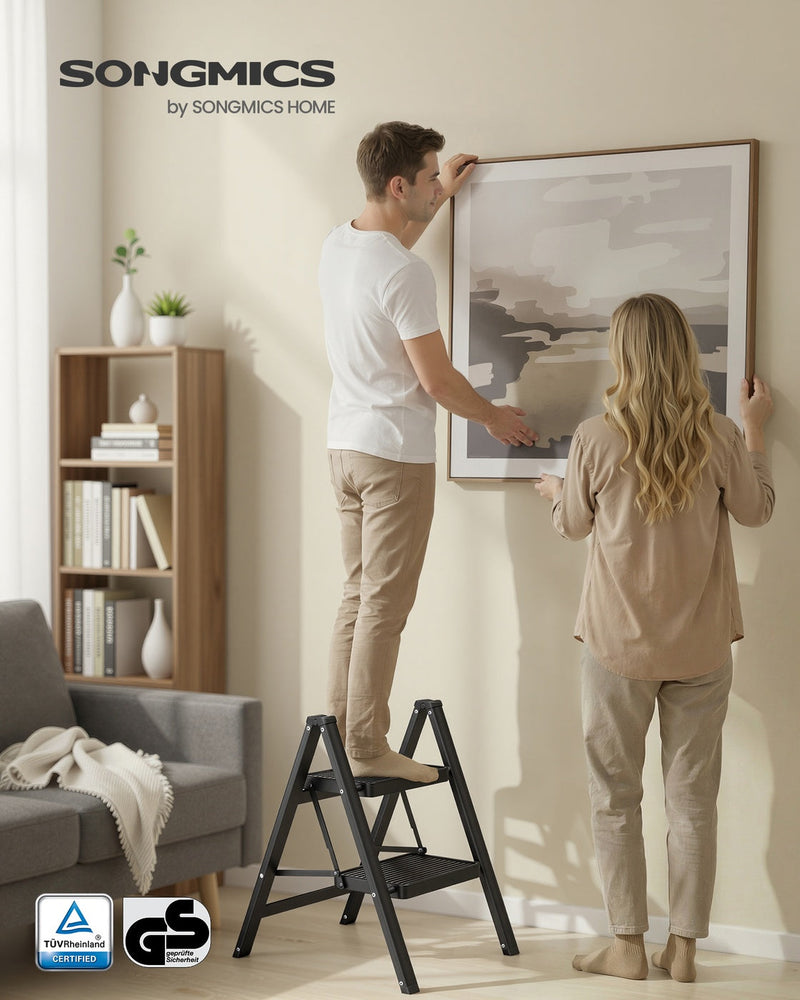 Couple hanging a framed picture with the black 2-step ladder open on a living room floor