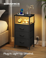 Ink black bedside table beside a bed with warm LED shelf lighting and a phone charging on top
