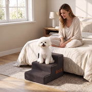 Small white dog sitting on the dog stairs beside a bed while a woman watches