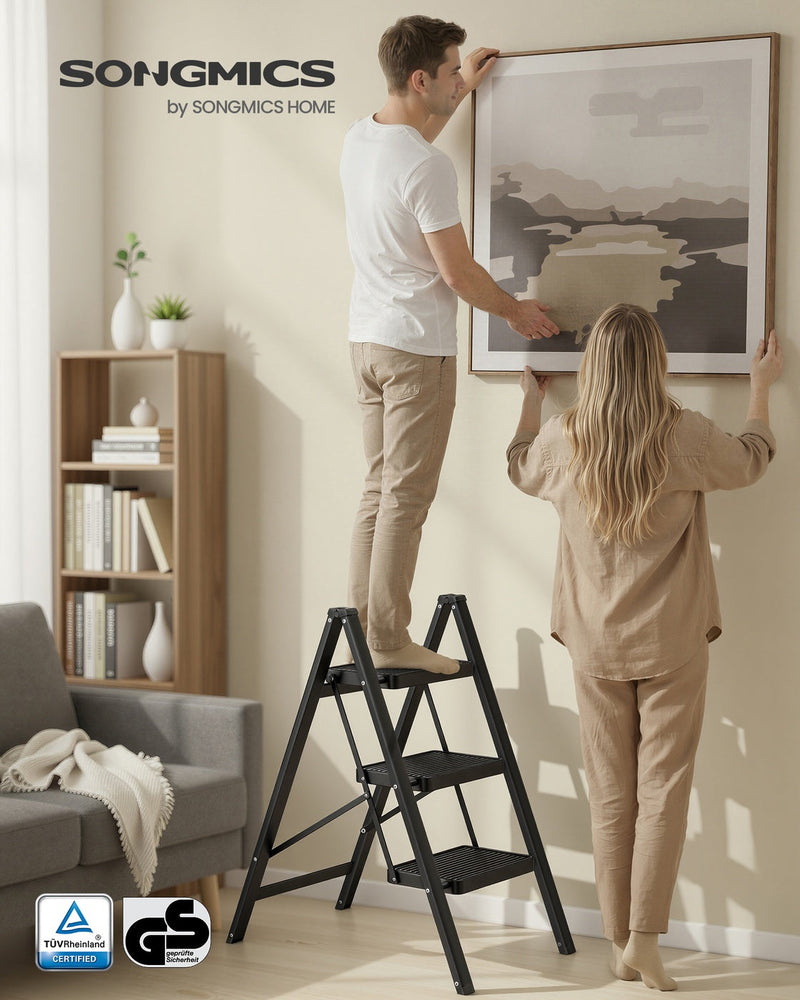 Two people hanging a picture with the black 3-step ladder open on a living room floor