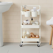 White storage trolley beside a basin holding towels, toiletries and a wicker tray in a tiled bathroom