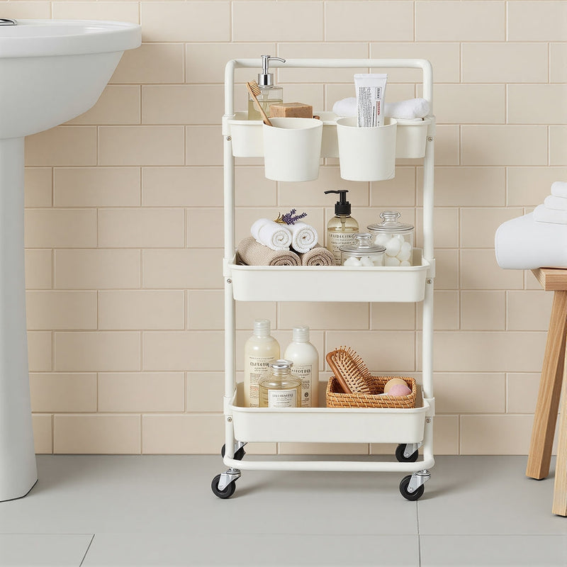 White storage trolley beside a basin holding towels, toiletries and a wicker tray in a tiled bathroom