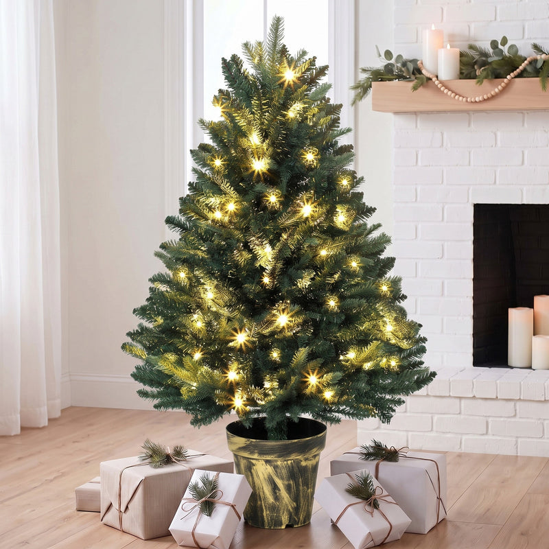 Lit Christmas tree in its pot base in front of a white brick fireplace with wrapped gifts