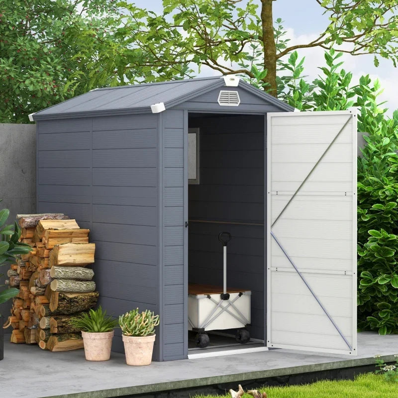 Grey plastic garden shed outdoors with the door open, logs and potted plants alongside