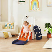 Toddler girl sliding down the orange slide in a living room with a rainbow wall decoration