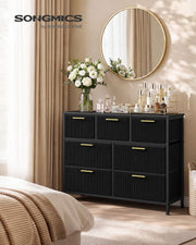 Black 7-drawer corduroy chest of drawers in a bedroom with flowers and a round mirror