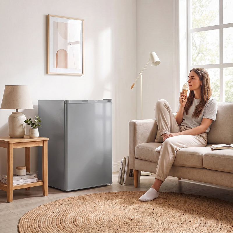 Grey mini freezer next to a sofa while a woman eats ice cream