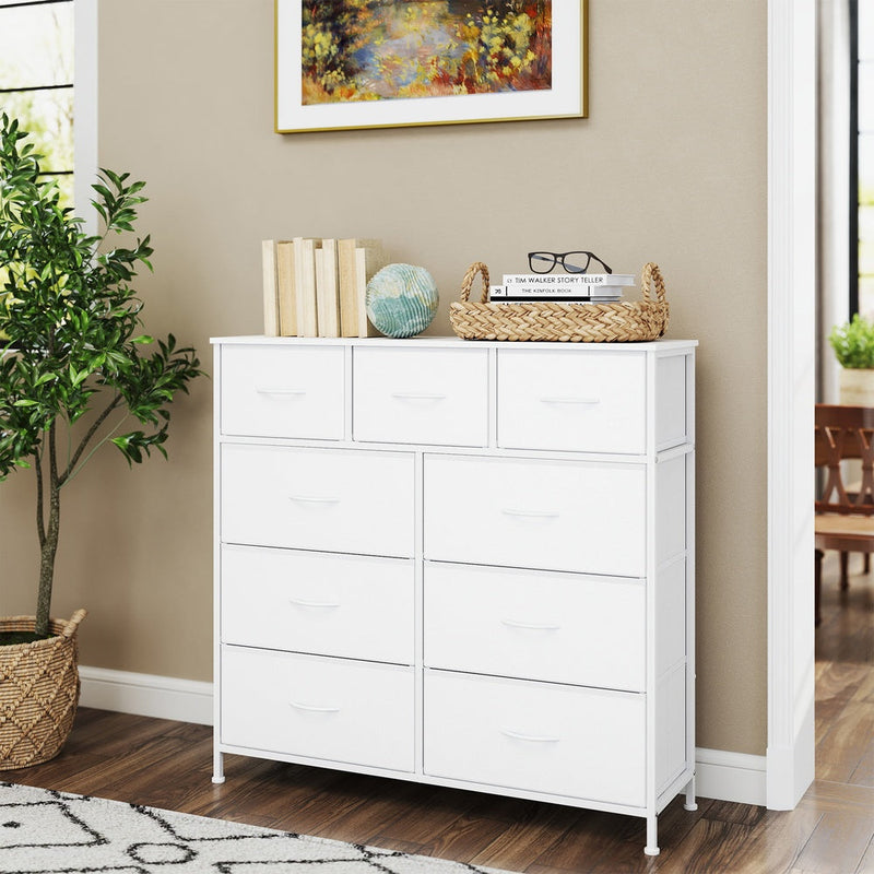 White chest of drawers in a hallway beside a potted tree, with books and a woven tray on top