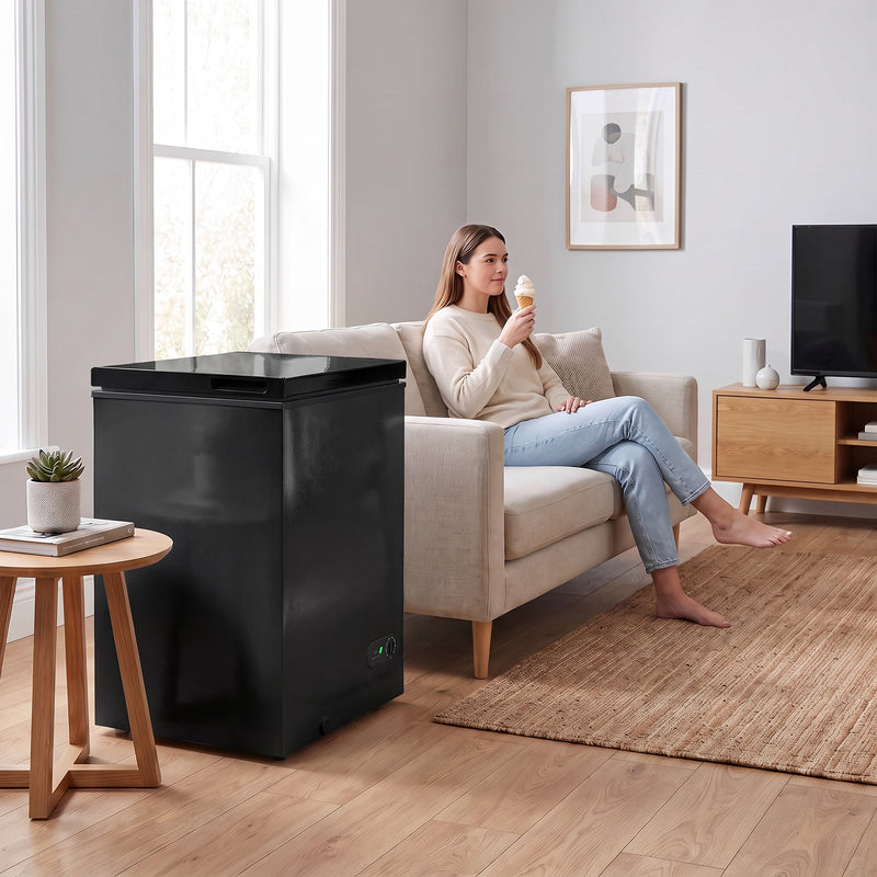 Black chest freezer in a living room while a woman eats ice cream on the sofa