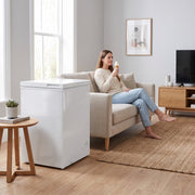 White chest freezer in a living room while a woman eats ice cream on the sofa