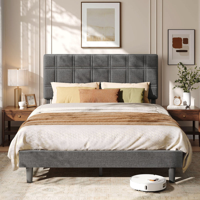 Upholstered double bed with grid-stitched headboard between two wooden bedside tables, robot vacuum below