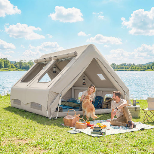 Two people with a dog sitting outside a large inflatable tent by a lake.