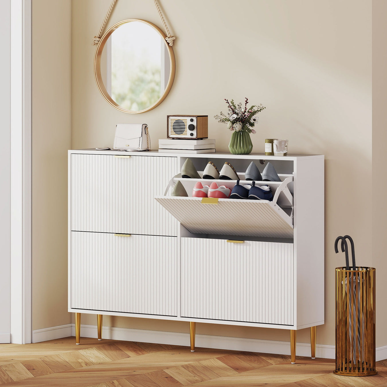 White shoe cabinet with fluted door fronts gold legs and handles with an open door revealing shoes, placed in a hallway with a round mirror and wooden floor.