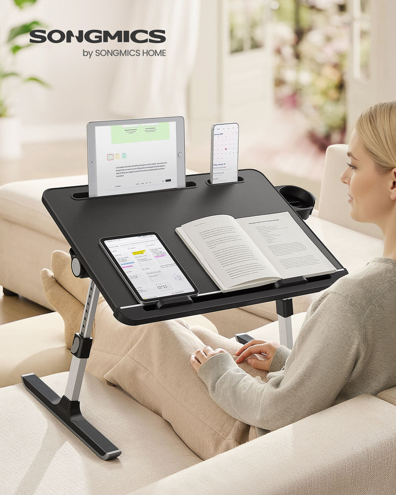 Woman on a sofa using the black laptop desk with a tablet, phone and open book