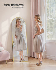 Woman in a grey dress checking her outfit in the arched floor mirror beside a pink armchair