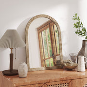 Brown arched mirror leaning on a wooden sideboard beside a table lamp