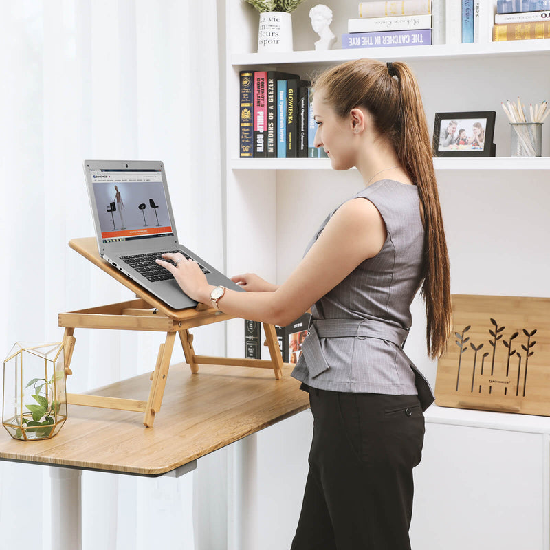 Woman standing at a desk typing on a laptop raised on the bamboo table in front of a bookshelf