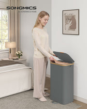 Woman opening a slate grey laundry hamper next to a bed