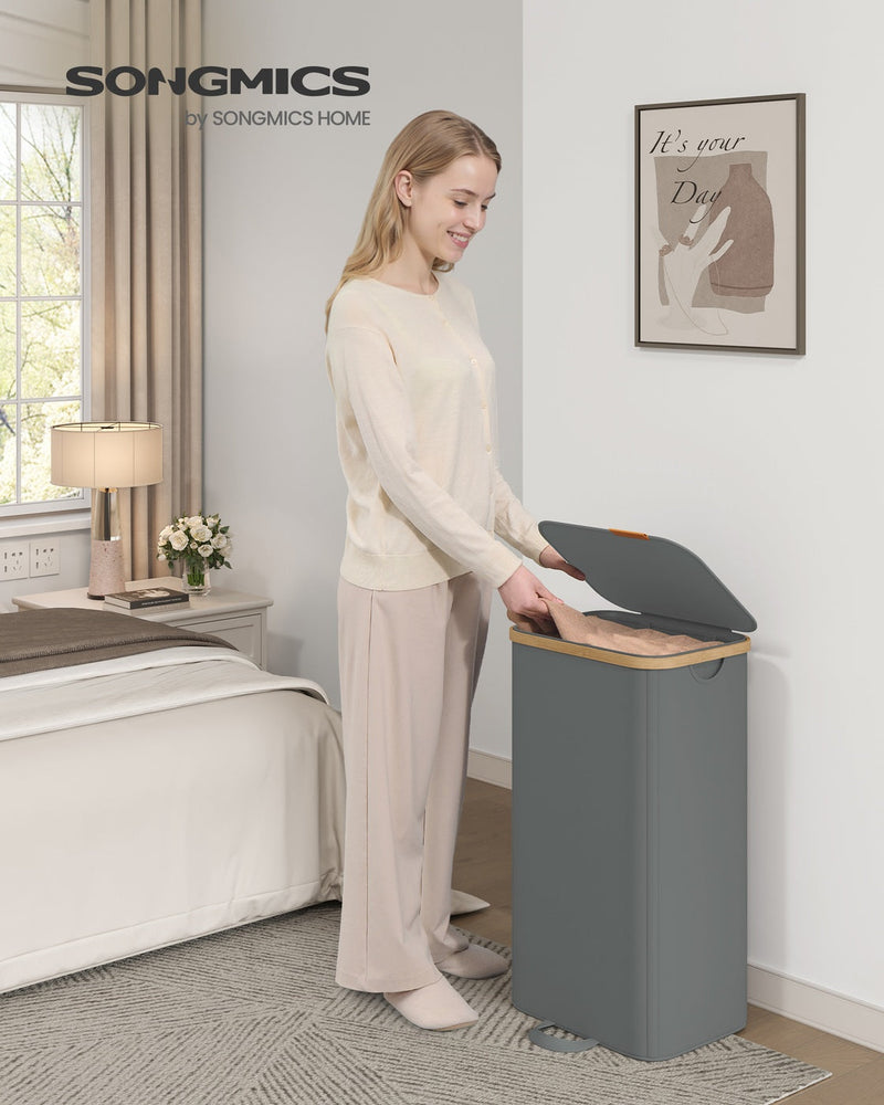 Woman opening a slate grey laundry hamper next to a bed