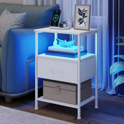 White bedside table in a living room with the shelf lit blue, a storage box on the lower shelf and a charging phone