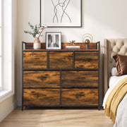 Rustic brown chest of drawers in a bedroom next to a bed with a plant and framed pictures on top