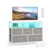 SONGMICS dove grey fabric 5-drawer chest of drawers styled as a TV stand with a wall-mounted TV and power outlet above.