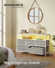 Dove grey fabric chest of drawers in a styled bedroom entryway beneath a round mirror, with one drawer open showing folded clothing under warm LED glow.