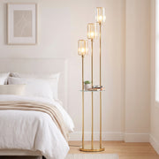 Gold tone floor lamp with glass shelf lit beside a bed in a bedroom