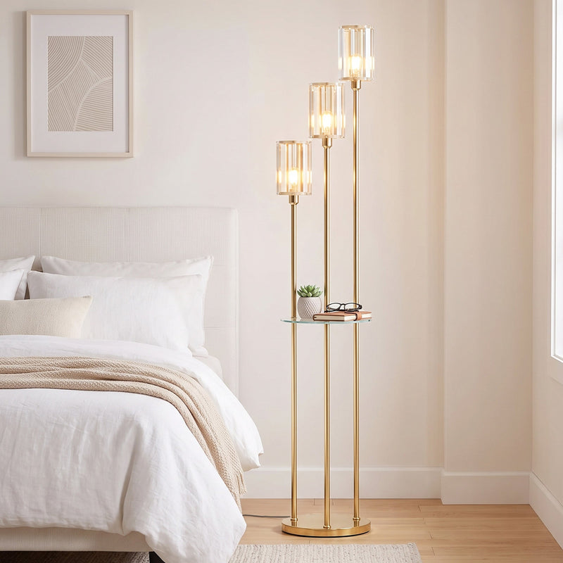 Gold tone floor lamp with glass shelf lit beside a bed in a bedroom