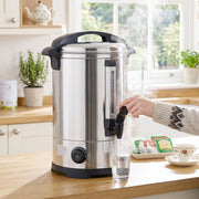 Hand pressing the tap of the 8L urn to fill a glass on a wooden kitchen worktop