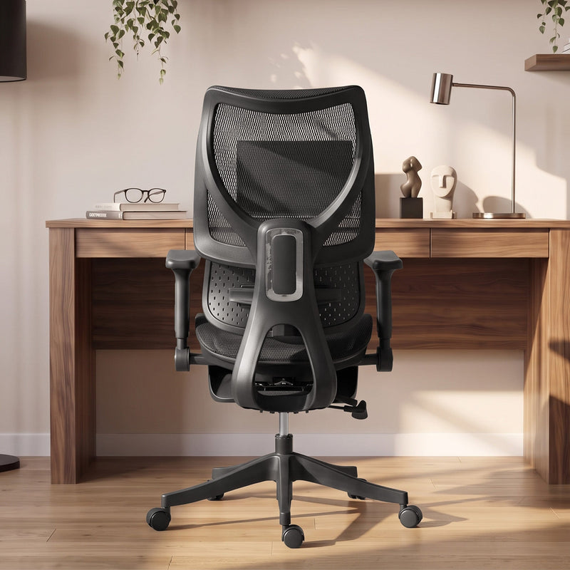 Black mesh office chair seen from behind facing a wooden desk with a lamp