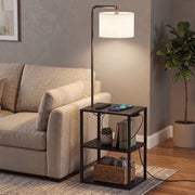 Black floor lamp with shelf table beside a beige sofa with a phone charging on the top shelf