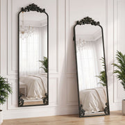 Two black baroque floor mirrors leaning against a bedroom wall reflecting a chandelier