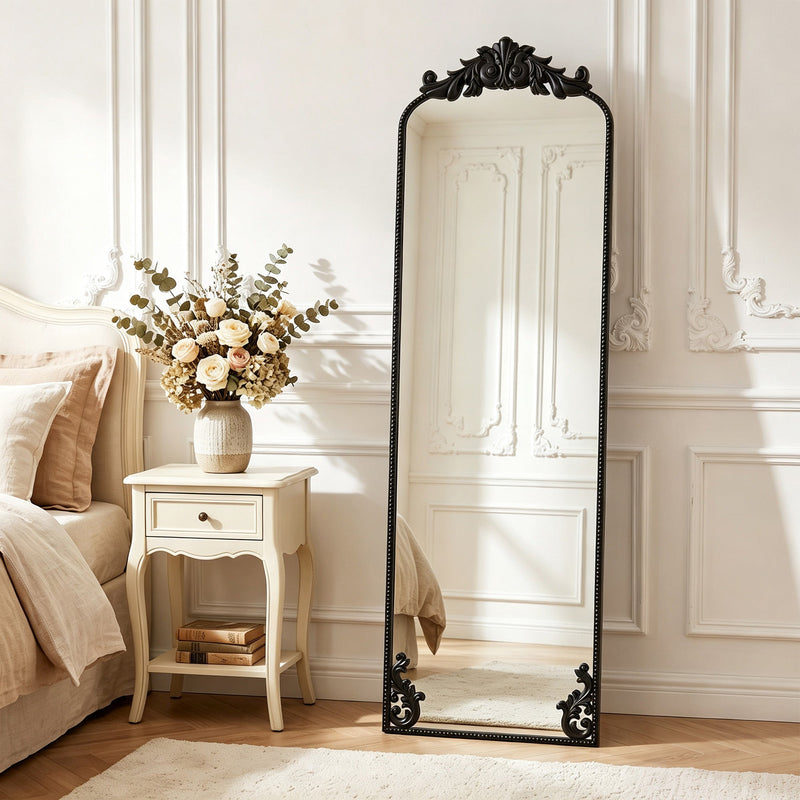Black baroque floor mirror leaning in a bedroom beside a nightstand with flowers