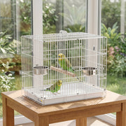 White bird travel cage on a wooden table by a garden window with two budgies inside