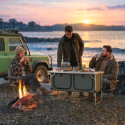 Group gathered around the camping cupboard on a beach at sunset with a kettle and mugs