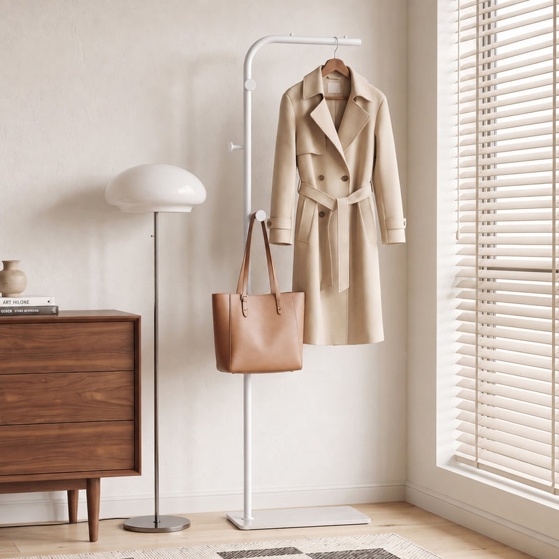 White clothes rail holding a trench coat and leather tote bag beside a wooden chest of drawers
