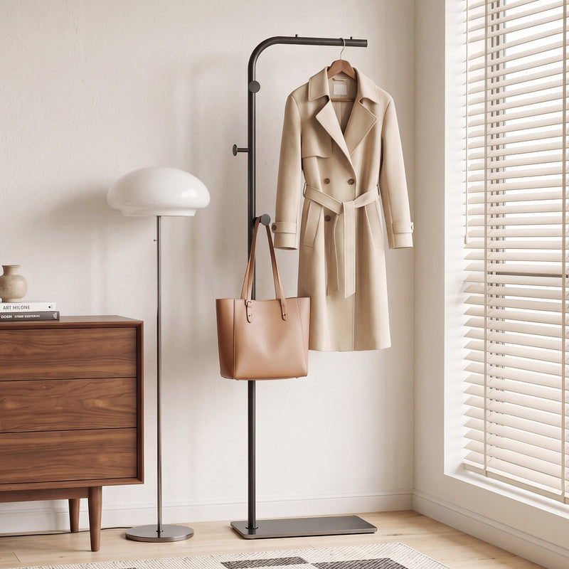 Black clothes rail holding a trench coat and leather tote bag beside a wooden chest of drawers