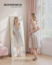 Woman checking her full-body reflection in the freestanding pale-gold mirror in a sunlit bedroom.