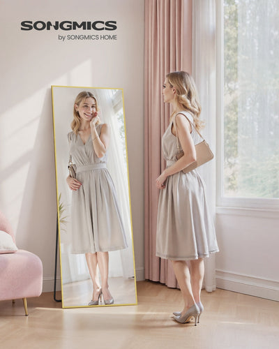 Woman checking her full-body reflection in the freestanding pale-gold mirror in a sunlit bedroom.