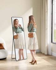 Woman checking her outfit in the black full-length mirror standing in a bright bedroom corner