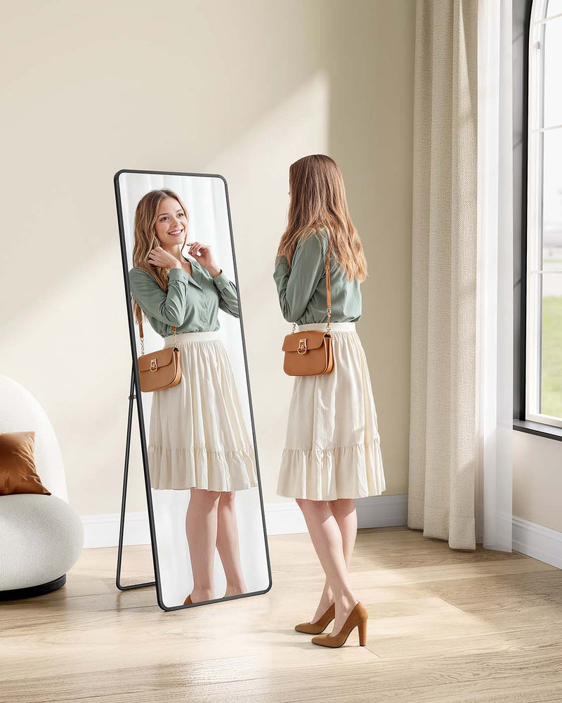 Woman checking her outfit in the black full-length mirror standing in a bright bedroom corner
