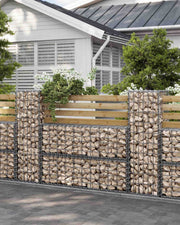 Stone-filled gabions used as a planted border beside a white house and shrubs