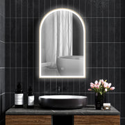 Lit arched mirror on a dark tiled wall above a black basin and tulips
