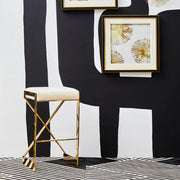 Ivory velvet bar stool with gold frame in front of a black and white backdrop with framed gold artwork