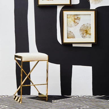 Ivory velvet bar stool with gold frame in front of a black and white backdrop with framed gold artwork