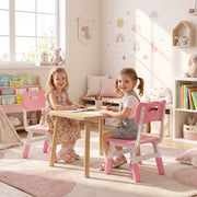 Two girls at a wooden table on kids chairs, with a bookcase and toys behind
