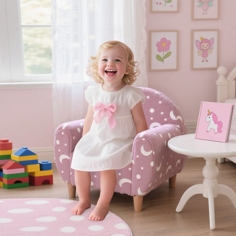 Toddler in a white dress sitting on the pink armchair beside a small play table
